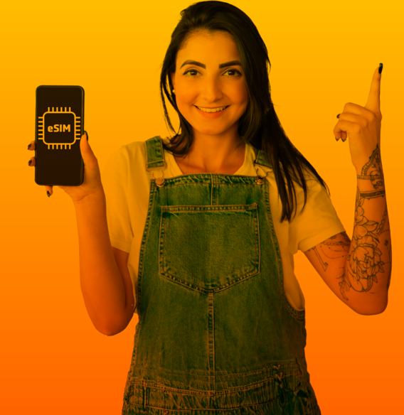 Junge, lächelnde Frau in Jeans-Latzhose hält ihr Handy mit eSIM-Karte hoch und zeigt mit der zweiten Hand nach oben.