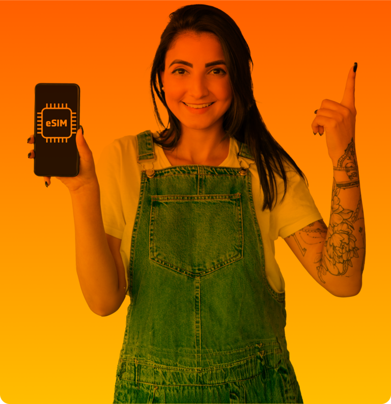 Junge, lächelnde Frau in Jeans-Latzhose hält ihr Handy mit eSIM-Karte hoch und zeigt mit der zweiten Hand nach oben.