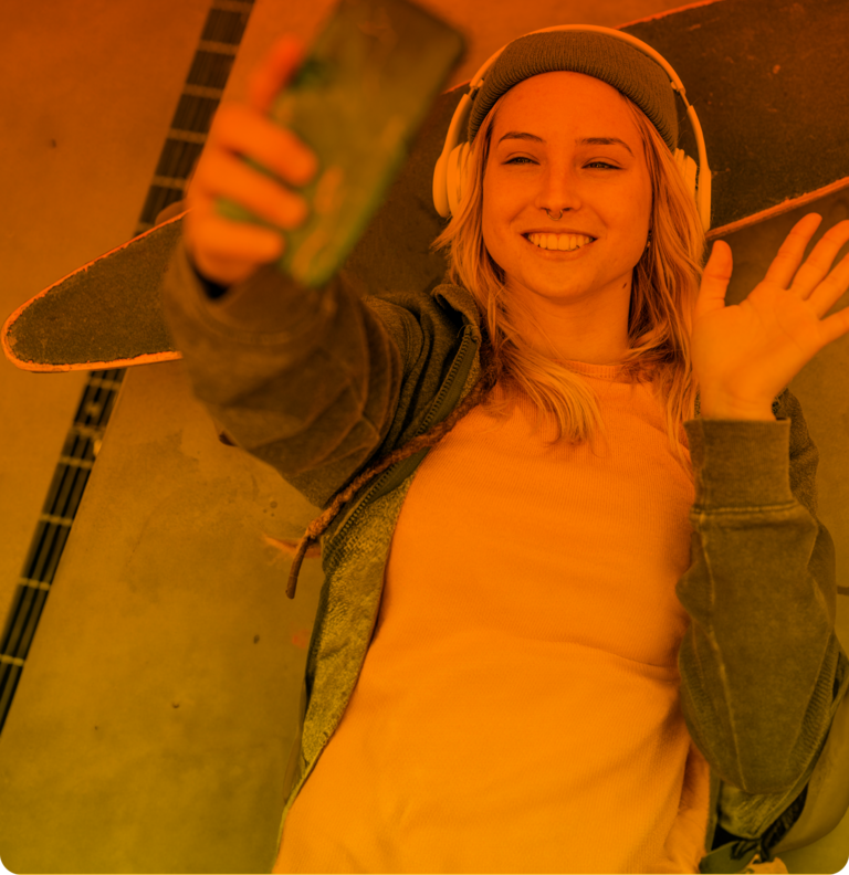 Die junge Frau mit Kopfhörern liegt winkend auf dem Skateboard und telefoniert online, dank ihres günstigen Handytarifs.